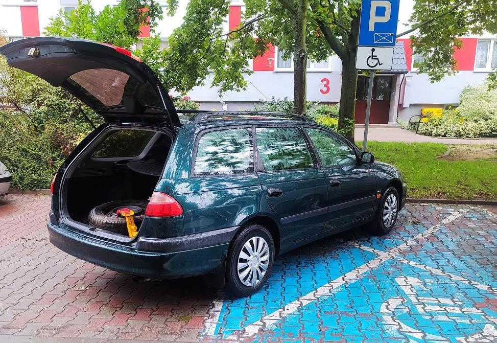 Samochód zaparkowany na miejscu dla osoby niepełnosprawnej. Otwarta klapa bagażnika, wewnątrz odkręcone koło z założonym urządzeniem blokującym koła. 